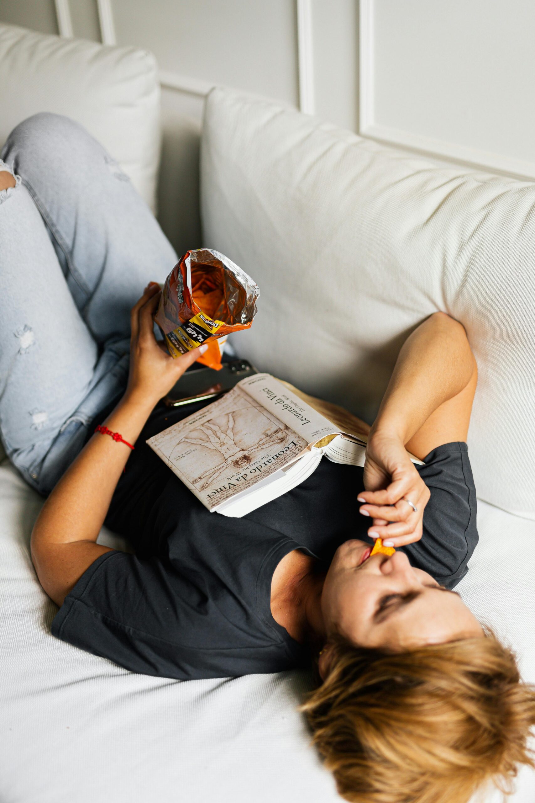 Woman lounging on a couch, eating chips, and reading a book in a casual setting.